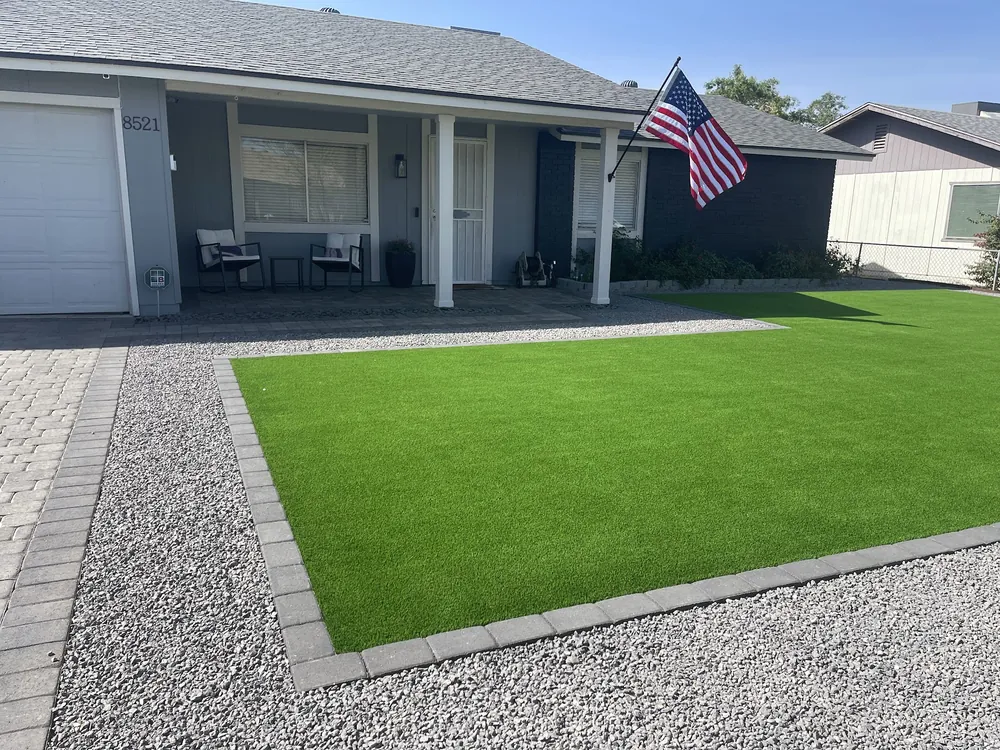 Front yard with a clean artificial turf lawn edged in pavers and gray gravel, in front of a home with a flag