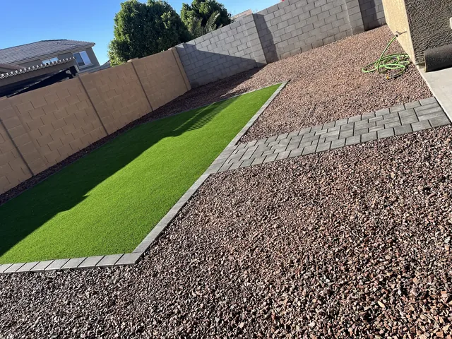 Artificial turf strip bordered by pavers beside a gravel bed along a block wall